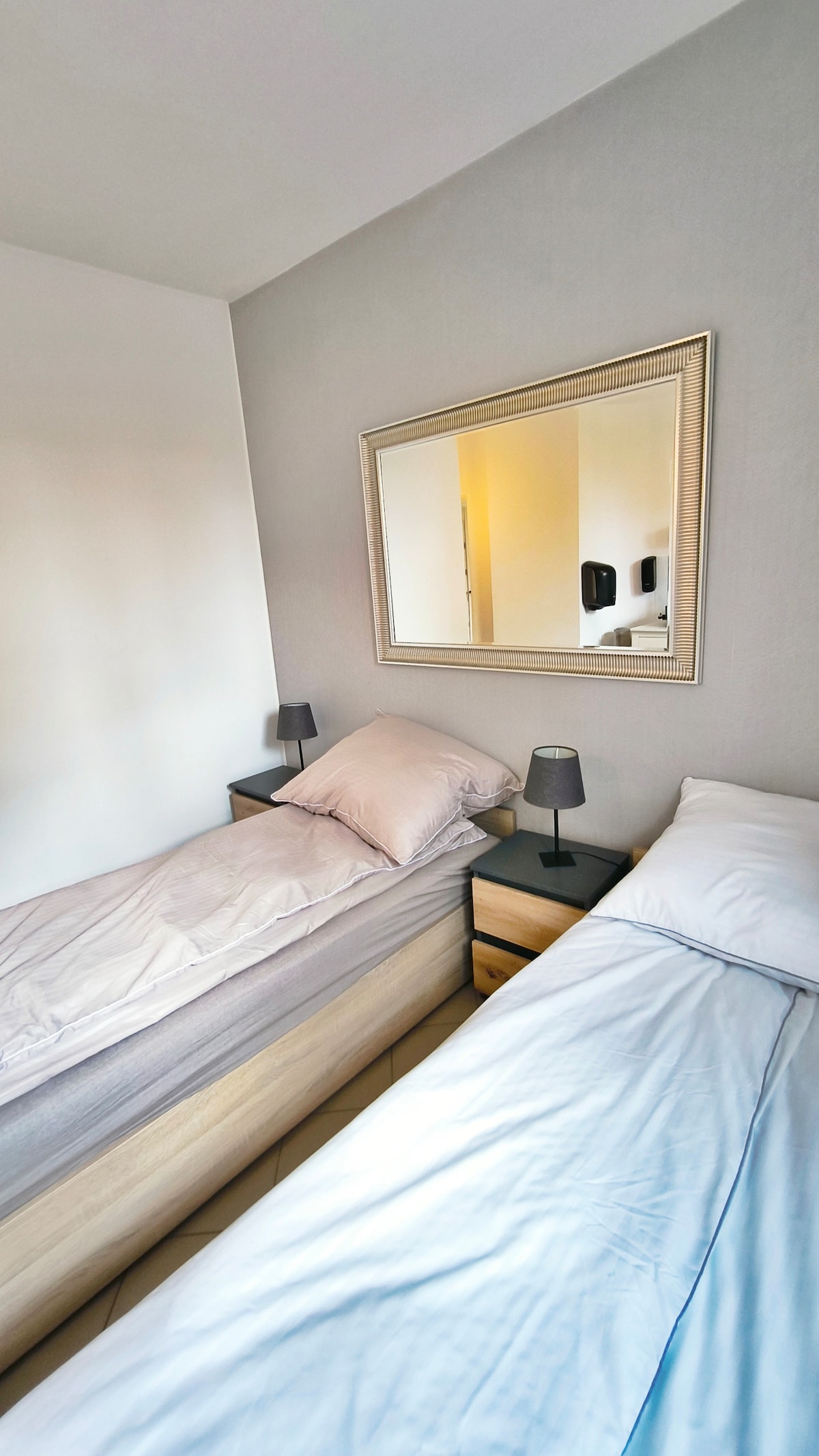The sleeping area features two single beds, each with soft bedding and placed beside compact nightstands. Small lamps are positioned on the nightstands, providing gentle illumination. A mirror hangs above the beds, reflecting the light from the nearby windows.