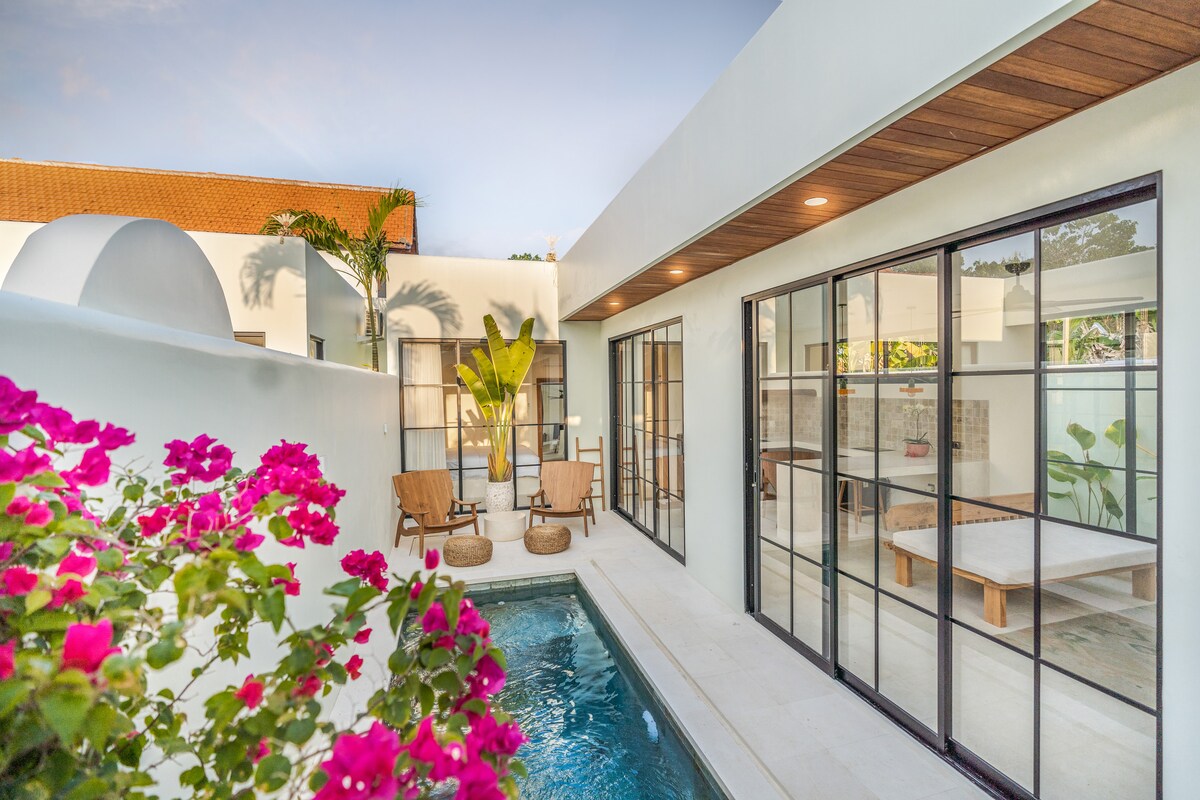 An open-air view of the villa reveals a private pool surrounded by minimalist wooden deck chairs and vibrant pink bougainvillea. Large glass doors enhance the seamless transition between indoor and outdoor spaces, with a modern design and tropical landscaping complementing the tranquil environment.