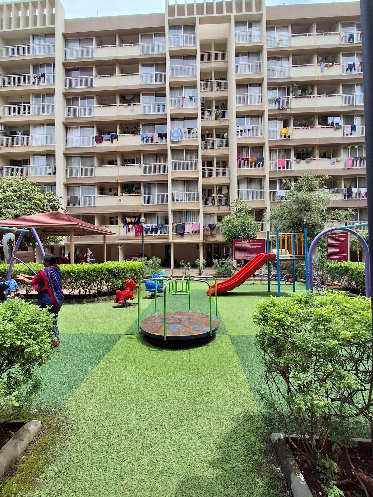 A vibrant playground is visible, featuring a spinning platform, a red slide, and climbing structures. Nearby, children are seen playing, while the backdrop includes a multi-story building with numerous balconies adorned with laundry. Green grass and shrubs frame the area, creating a welcoming outdoor environment.