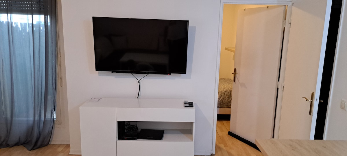 A flat-screen television is mounted on the wall above a sleek white media console. The console features an open shelf, housing electronic devices. A door leads to a softly lit bedroom, while sheer gray curtains cover a window, allowing natural light to brighten the room.