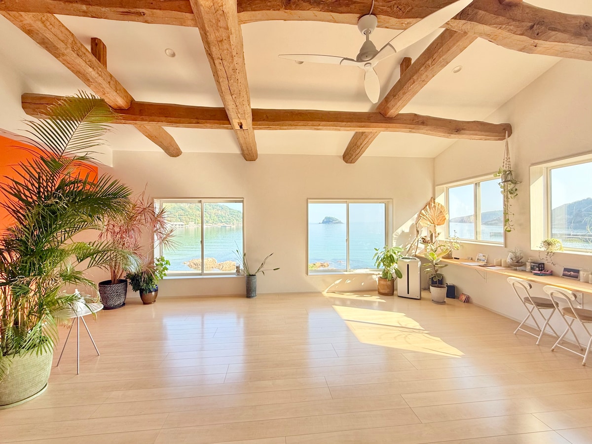A spacious living area features high ceilings supported by wooden beams. Large windows provide a panoramic view of the sea, while potted plants add greenery. A small table with two chairs is positioned thoughtfully near the window, making it a perfect spot for reflection.