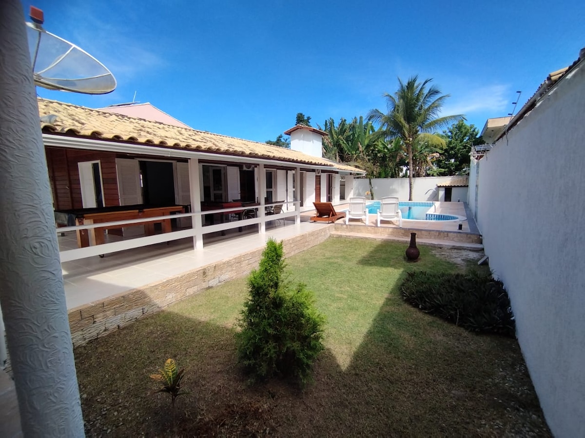A spacious outdoor area features a well-maintained lawn and a refreshing blue pool. The main building, with a tiled roof, is surrounded by several shaded seating areas. Lush greenery and palm trees add a natural touch, creating a relaxing environment.