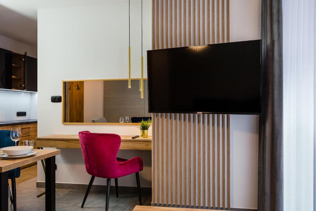 A modern living area features a wall-mounted flat-screen TV above a wooden console. A round dining table is set for two, accompanied by a vibrant pink chair. Natural light streams in through sheer curtains, illuminating the neutral color palette of the space.