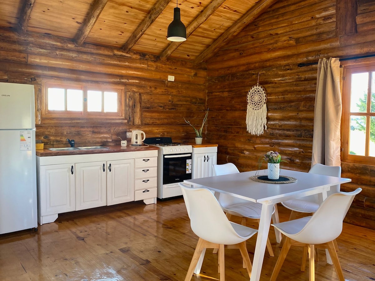 A rustic kitchen area features wooden walls and a high ceiling with exposed beams. A white dining table surrounded by four chairs is at the center. Kitchen appliances, including a refrigerator, stovetop, and kettle, are neatly arranged, providing a functional space for cooking and dining.