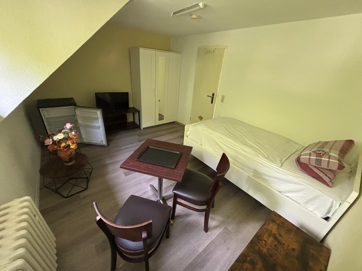 A well-organized room features a single bed with an unmade white bedding. A small table with two chairs is positioned in the center. A cabinet and a television are present, along with a refrigerator. A decorative flower arrangement adds a subtle touch.