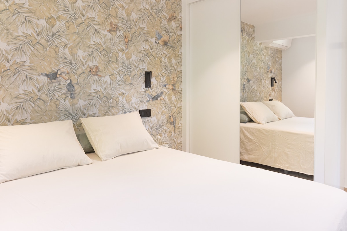 The sleeping area features a spacious bed adorned with soft, neutral bedding. A decorative wallpaper with a nature-inspired pattern complements the room's calm ambiance. A mirror reflects the inviting setting, enhancing the sense of space and light.