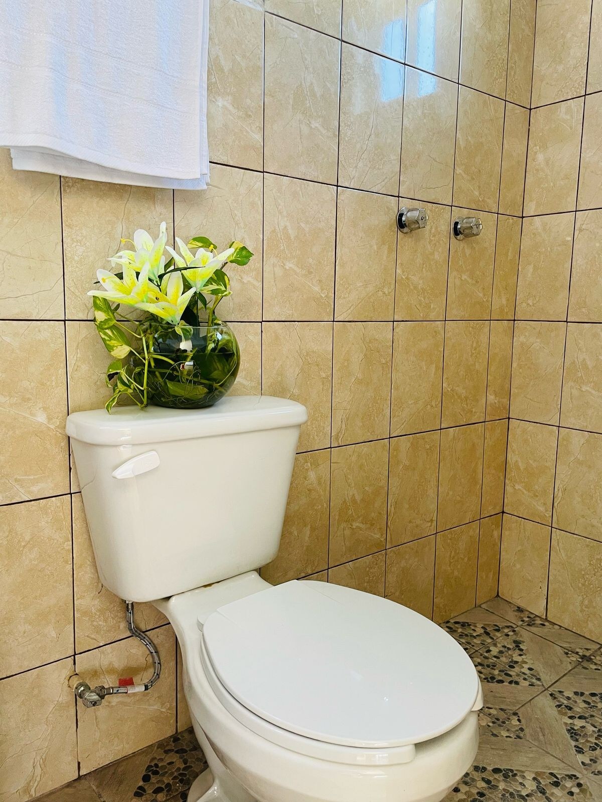 A well-maintained bathroom features beige tiled walls and flooring. A white toilet is positioned against the wall, topped with a vase containing fresh flowers. Natural light filters in, enhancing the clean and simple aesthetic of the space.