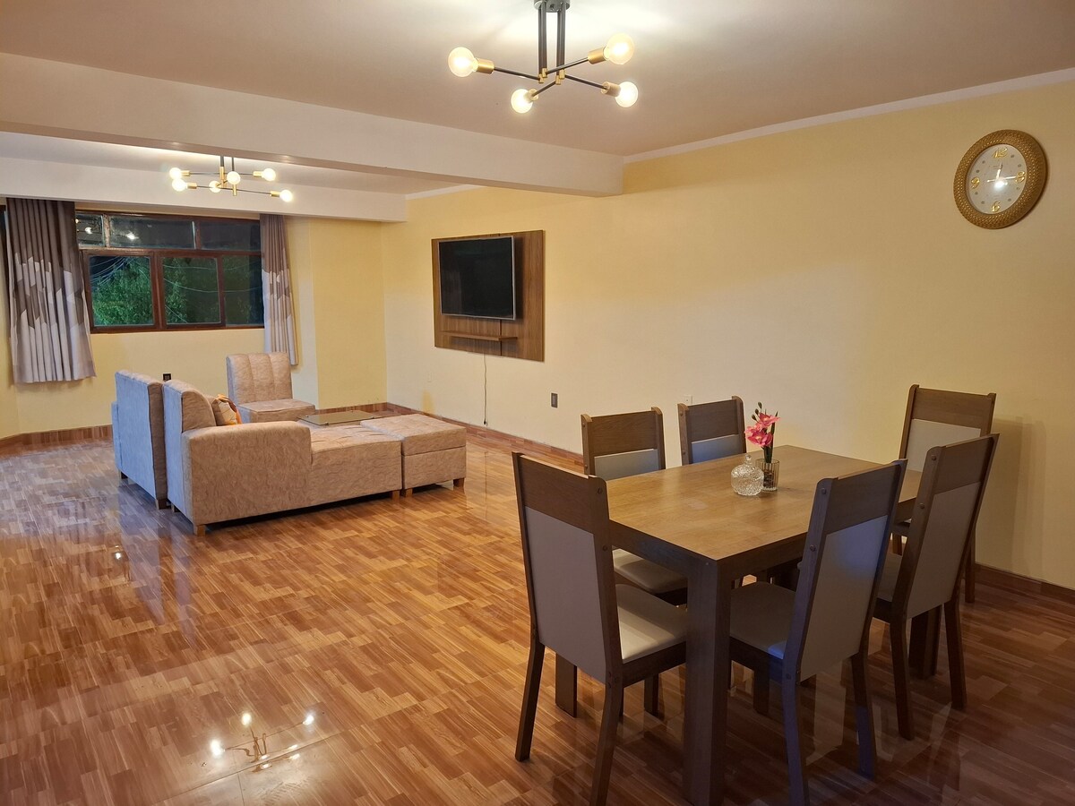 An inviting living area features a comfortable sofa facing a wall-mounted television. A dining table with six chairs is positioned nearby, with natural light filtering through large windows. Warm wood floors are present throughout, complemented by soft yellow walls and modern light fixtures.