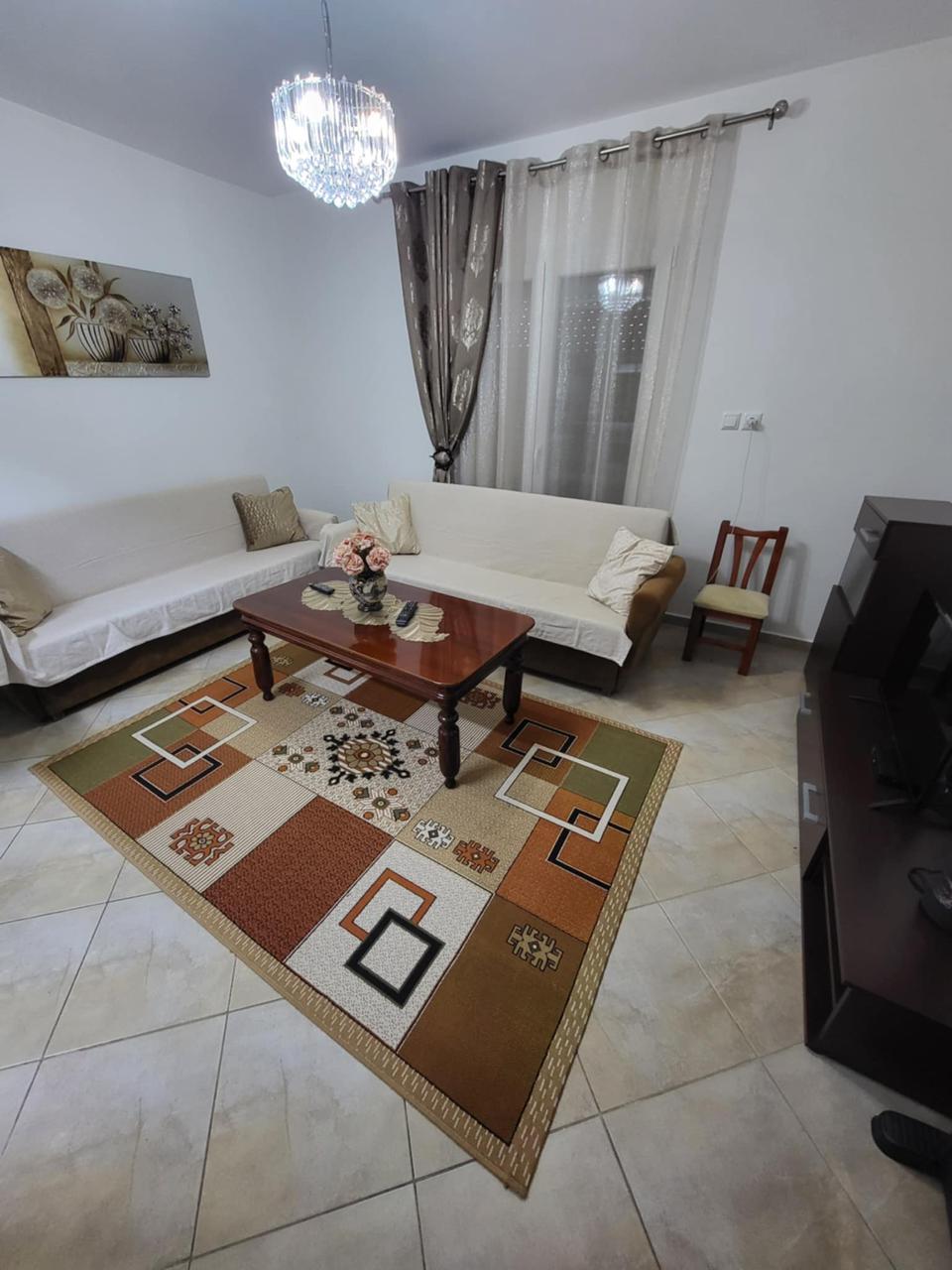 A welcoming living area features a hardwood table at the center, surrounded by two sofa beds draped in neutral covers. A decorative area rug adds warmth, while a small chair is positioned nearby. Natural light filters through a sheer curtain, complementing the room's soft color palette.