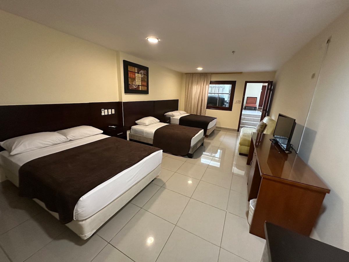 The spacious room features two beds with dark brown coverings, along with a television on a wooden stand. A large window allows natural light to enter, while a cozy armchair sits in one corner. The tiled floor provides a clean and modern appearance.