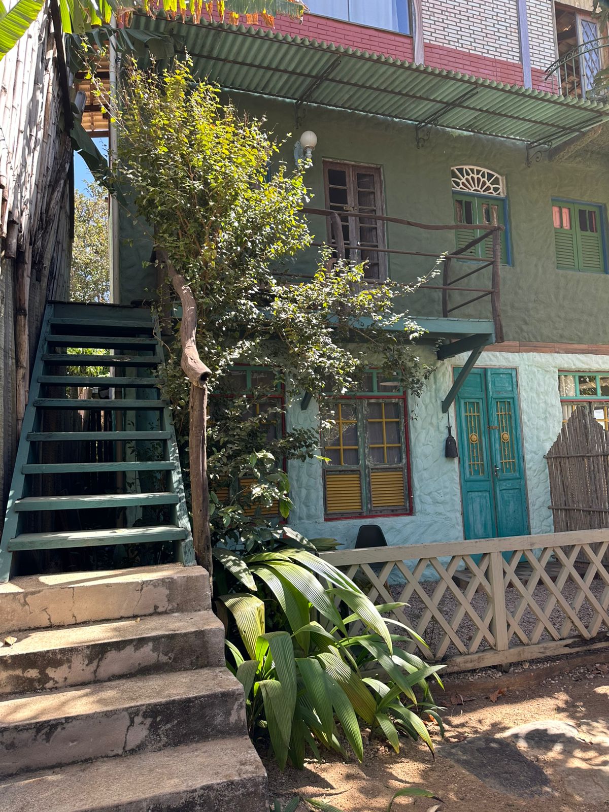 A vibrant exterior showcases a two-story building with green and blue accents. A staircase leads to the upper level, surrounded by lush greenery. Colorful windows and a decorative balcony emphasize the building's architectural charm, merging nature with artistic elements.