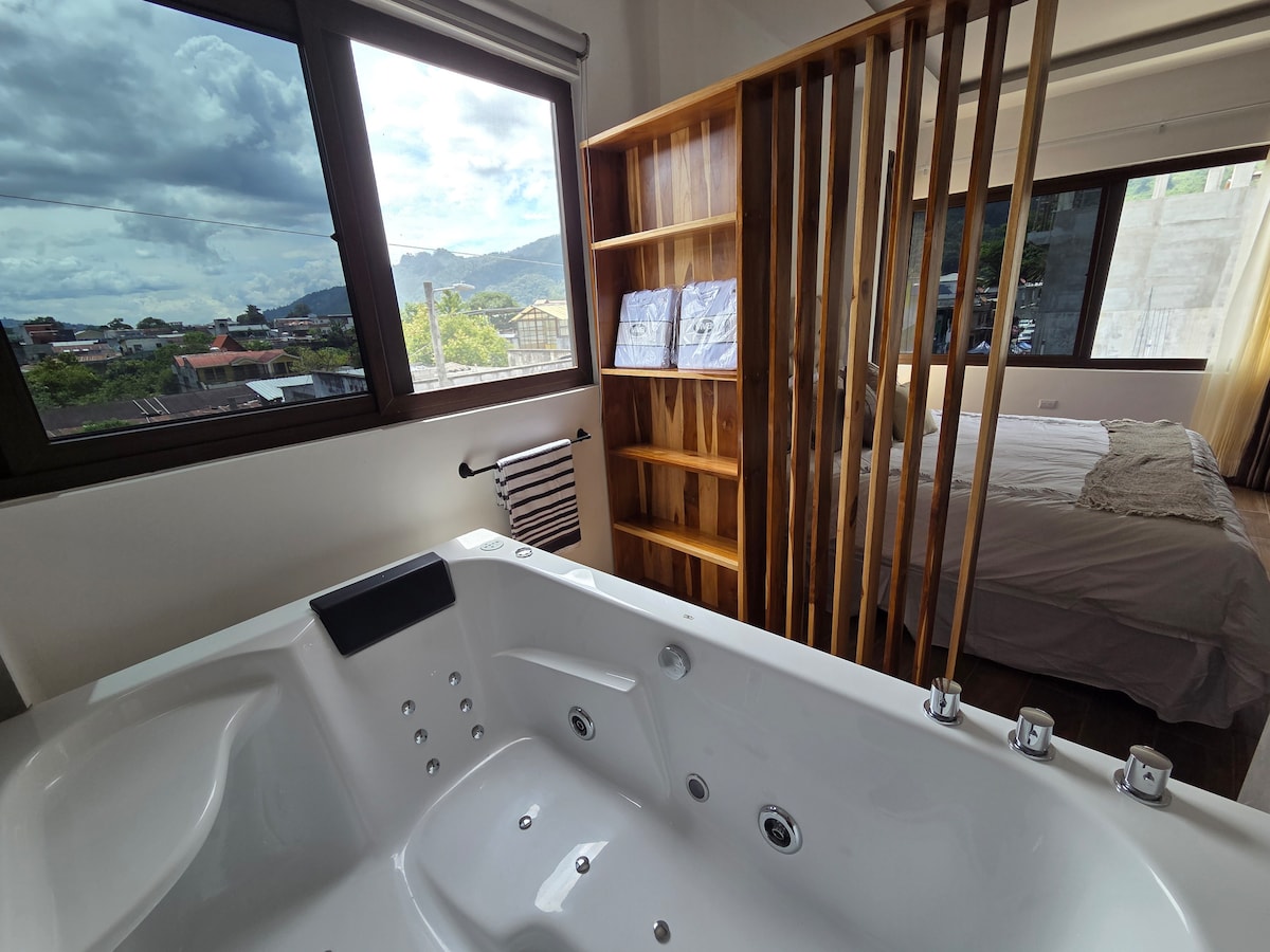 A modern bathroom features a spacious hydromassage tub positioned near a large window, offering views of the surrounding landscape. A wooden divider allows for a sense of privacy while showcasing a serene sleeping area with natural light flowing through the nearby window.