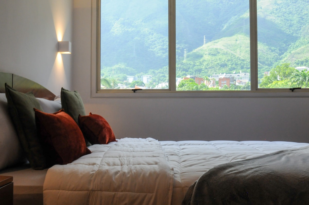 A spacious bedroom is highlighted by a large window, offering a view of lush mountains. A comfortable bed is dressed in soft linens, complemented by decorative pillows in warm tones. Natural light fills the room, enhancing the serene environment.