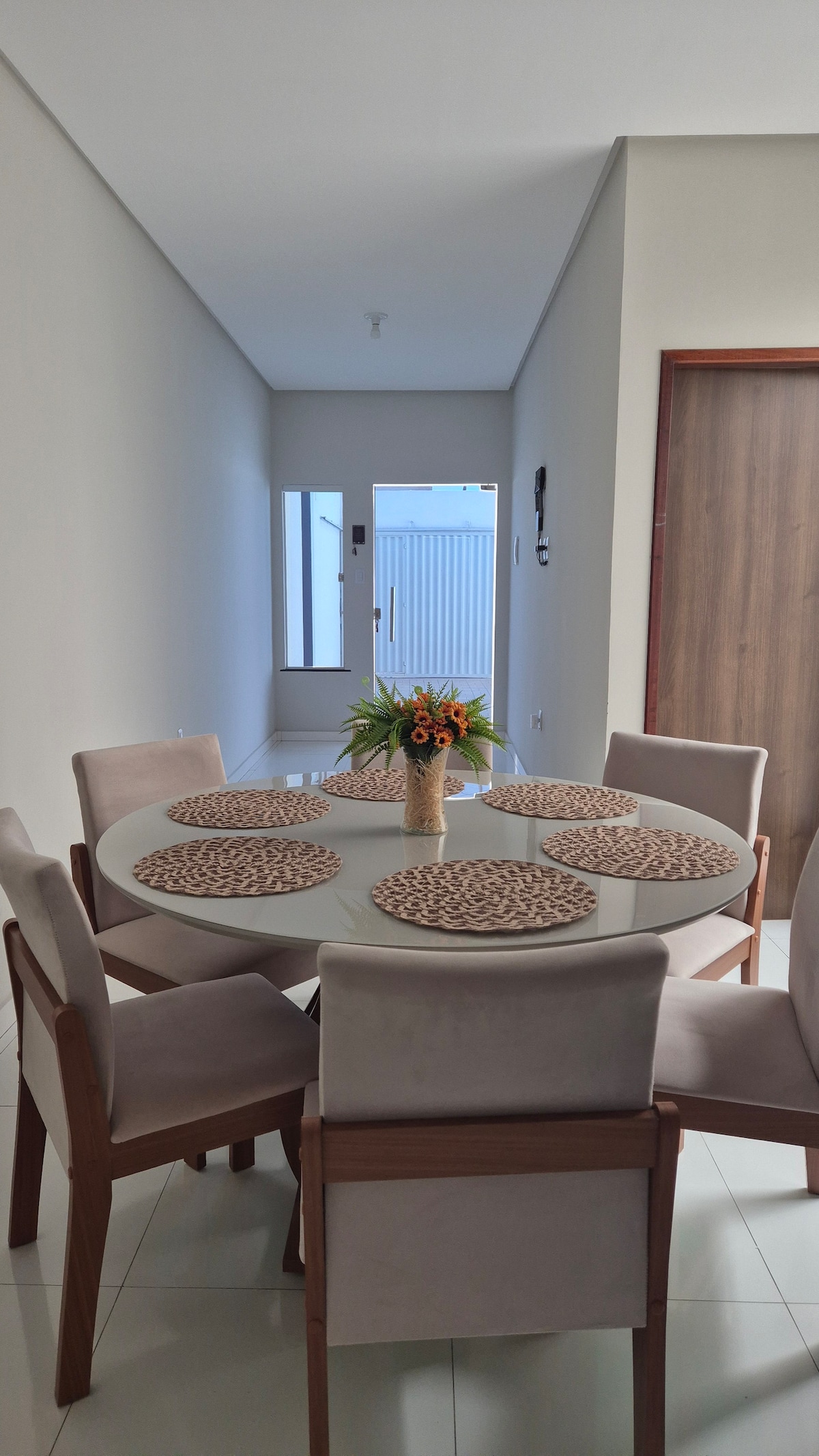A round dining table is set with six beige placemats, each accompanied by a small flower vase at the center. Surrounding the table are wooden chairs with soft upholstery. A faint view of a hallway and an exterior door can be seen in the background.
