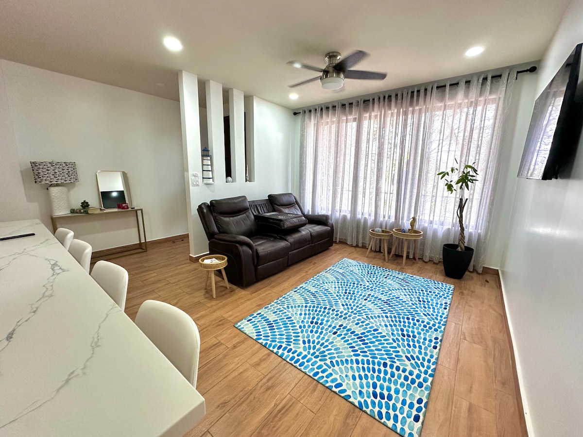 A living area features a dark brown leather couch positioned opposite a wall-mounted television. Natural light enters through large windows dressed in sheer white curtains. A decorative plant and a small side table are positioned near a patterned blue and white rug on the wooden floor.