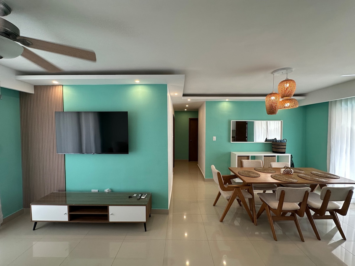 A modern living area features a large flat-screen television mounted to the wall, complemented by a wooden console. A stylish dining table with six chairs occupies the space, alongside warm, woven pendant lighting. Light reflects off the shiny tiled floor, creating an airy ambiance.