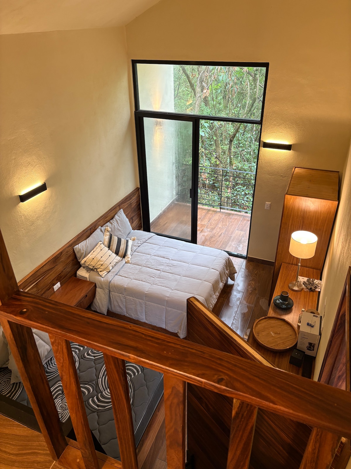 A bright sleeping area is visible from an elevated view, featuring a spacious bed with neutral bedding and accent pillows. Large glass doors invite natural light and offer views of surrounding greenery. Wooden furnishings complement the warm tones of the room.