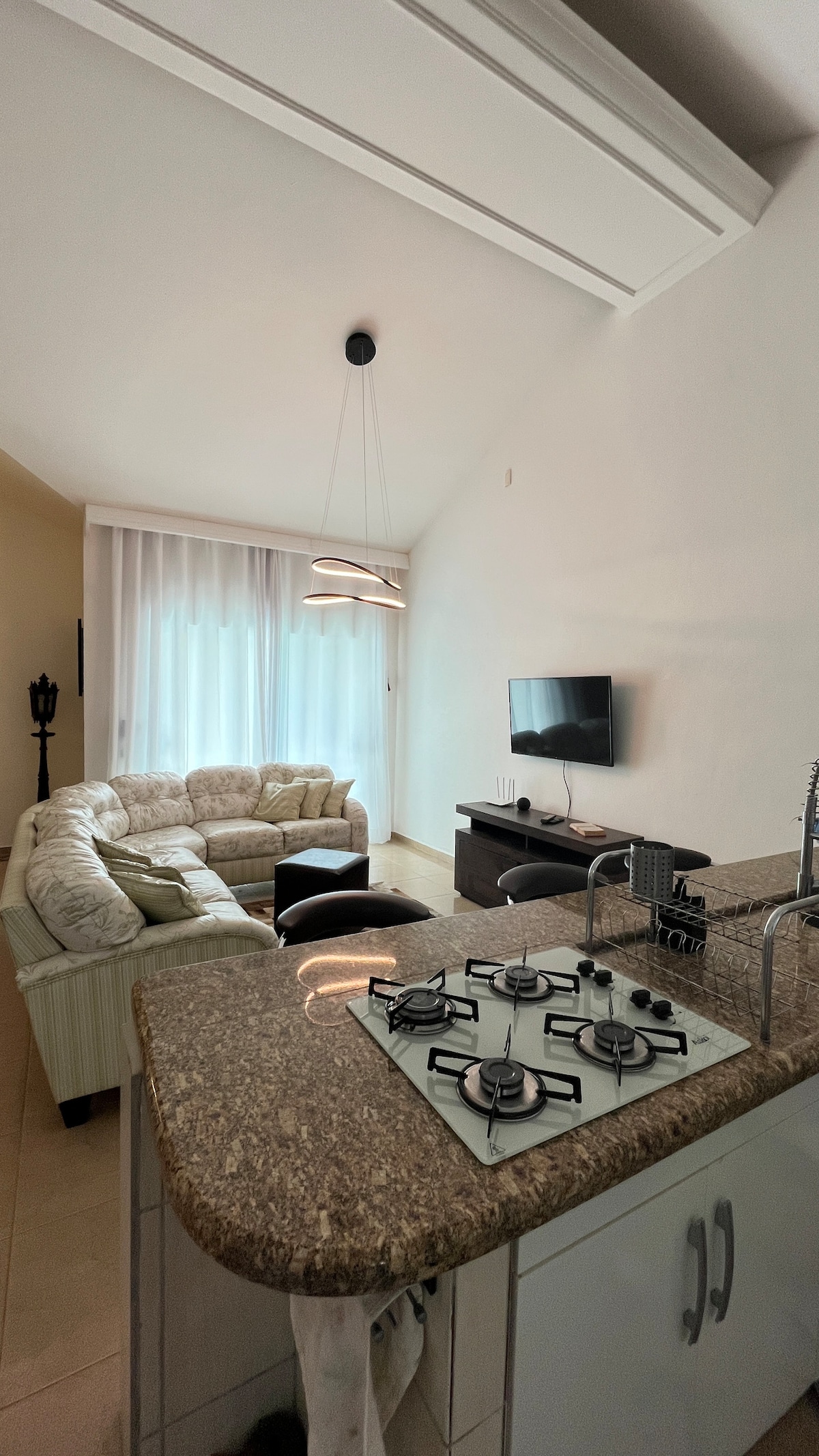 A living area features a light-colored sectional sofa and a sleek coffee table. Natural light filters through large windows with sheer curtains. A wall-mounted television is visible, alongside a kitchen counter equipped with a gas stove, enhancing the open and functional layout.