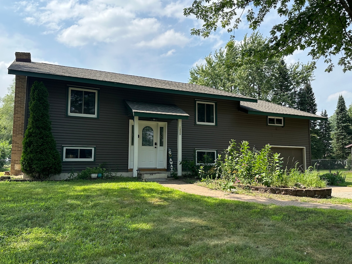 A welcoming exterior view of a two-story house, featuring a combination of dark siding and a light front door. A paved pathway leads towards the entry, bordered by well-maintained grass and a garden with tall plants and flowers, under a partly cloudy sky.