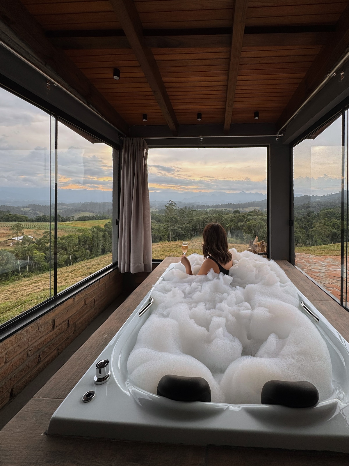 A modern bath area features a spacious tub filled with bubbles, positioned in front of large glass windows. Expansive views of the surrounding hills and sky can be seen through the windows, enhancing the sense of relaxation in the space.