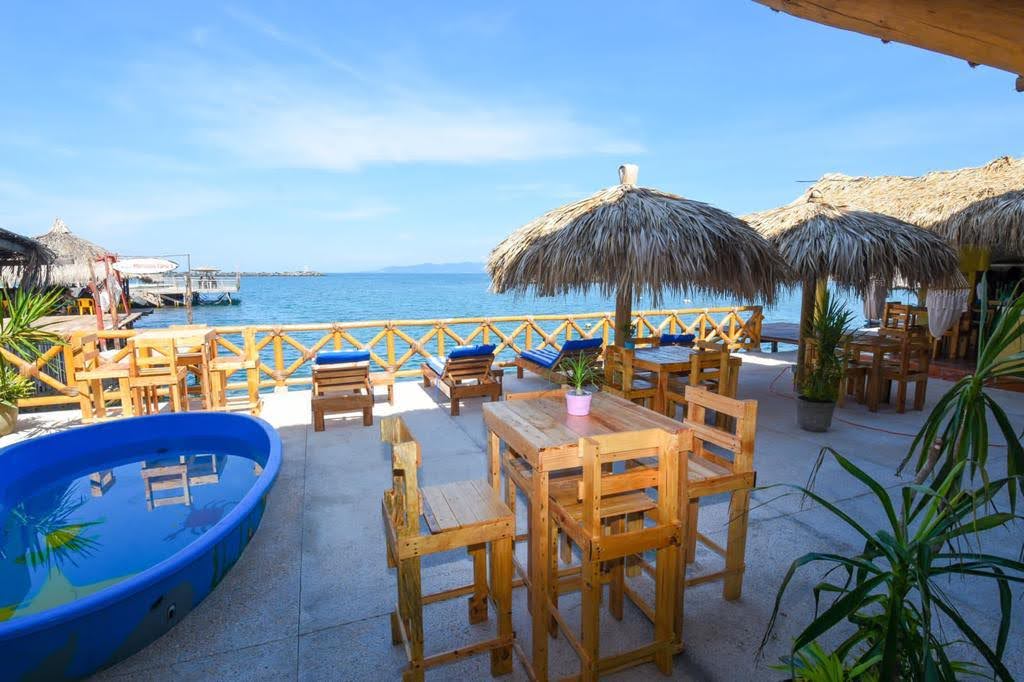 An outdoor dining area features wooden tables and chairs under straw umbrellas. A small pool is positioned nearby, with the sea visible in the background. Lush tropical plants are included in the setting, contributing to a relaxed coastal ambiance.
