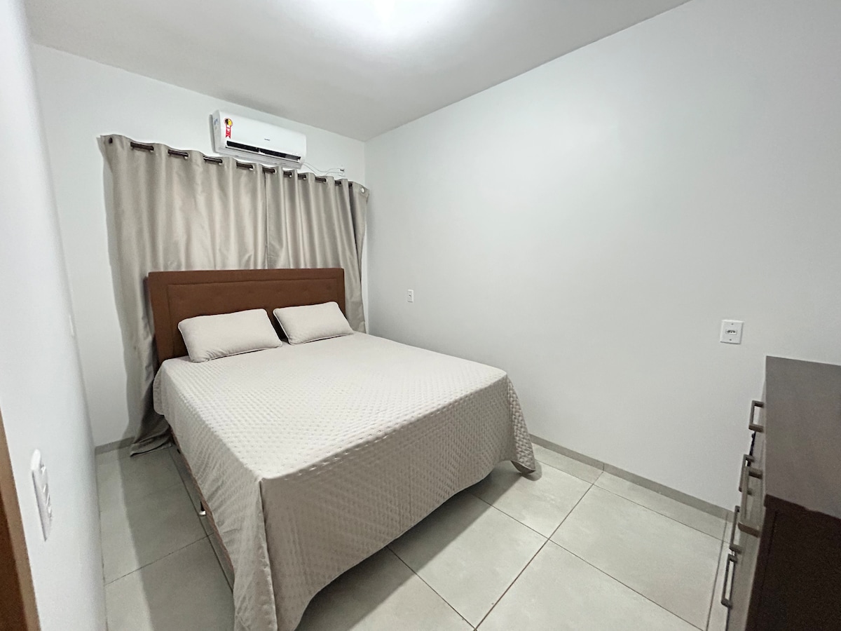 A well-ventilated bedroom presents a spacious bed with neutral bedding. Soft, muted curtains frame the window, allowing for natural light. A dresser is positioned against the wall, completing the room's clean and simple design.