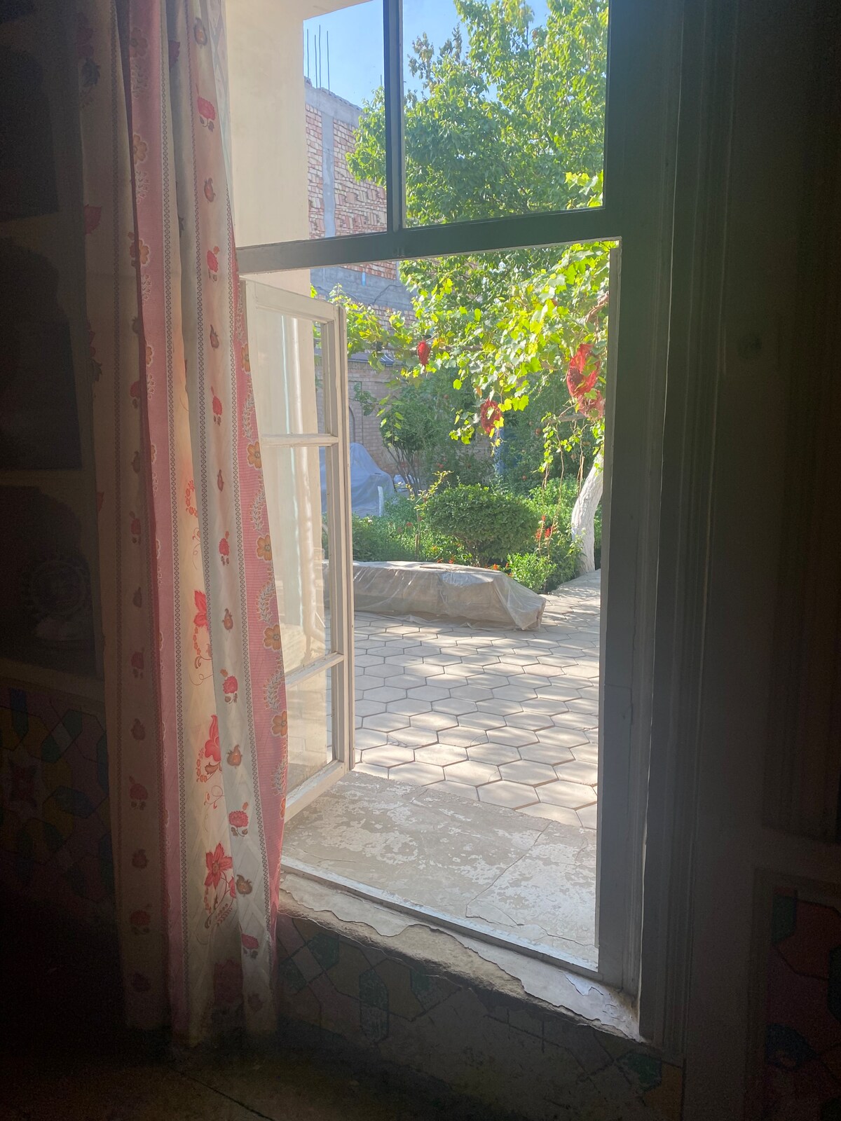 A window with white frames and floral curtains opens to a private courtyard. Soft sunlight illuminates the outdoor space, revealing greenery and a stone path. A partially covered seating area is visible beyond the entrance, inviting a connection with nature.