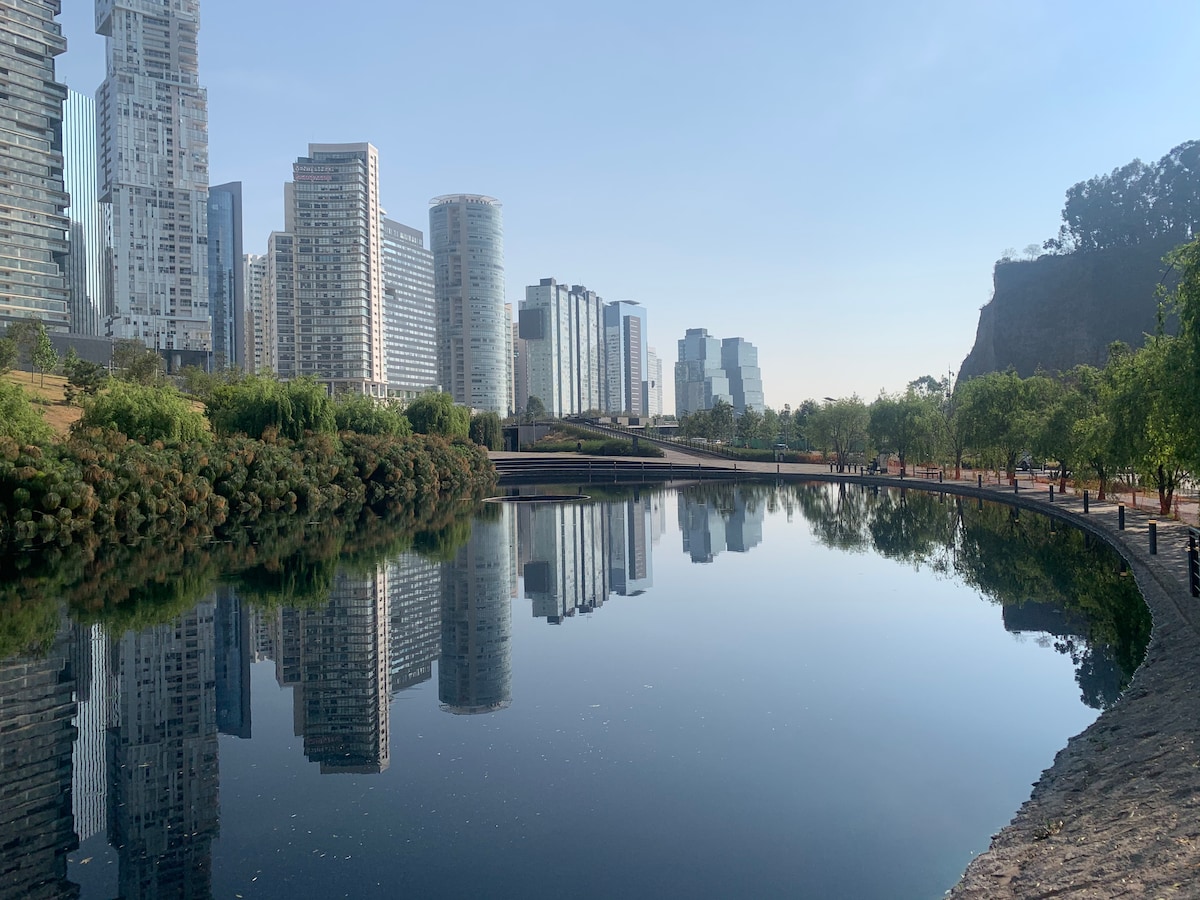A calm lake reflects the modern skyline, showcasing a blend of glass and concrete buildings. Lush trees line the walkway around the water, providing greenery against the backdrop of urban architecture. The serene atmosphere is enhanced by the clear sky.