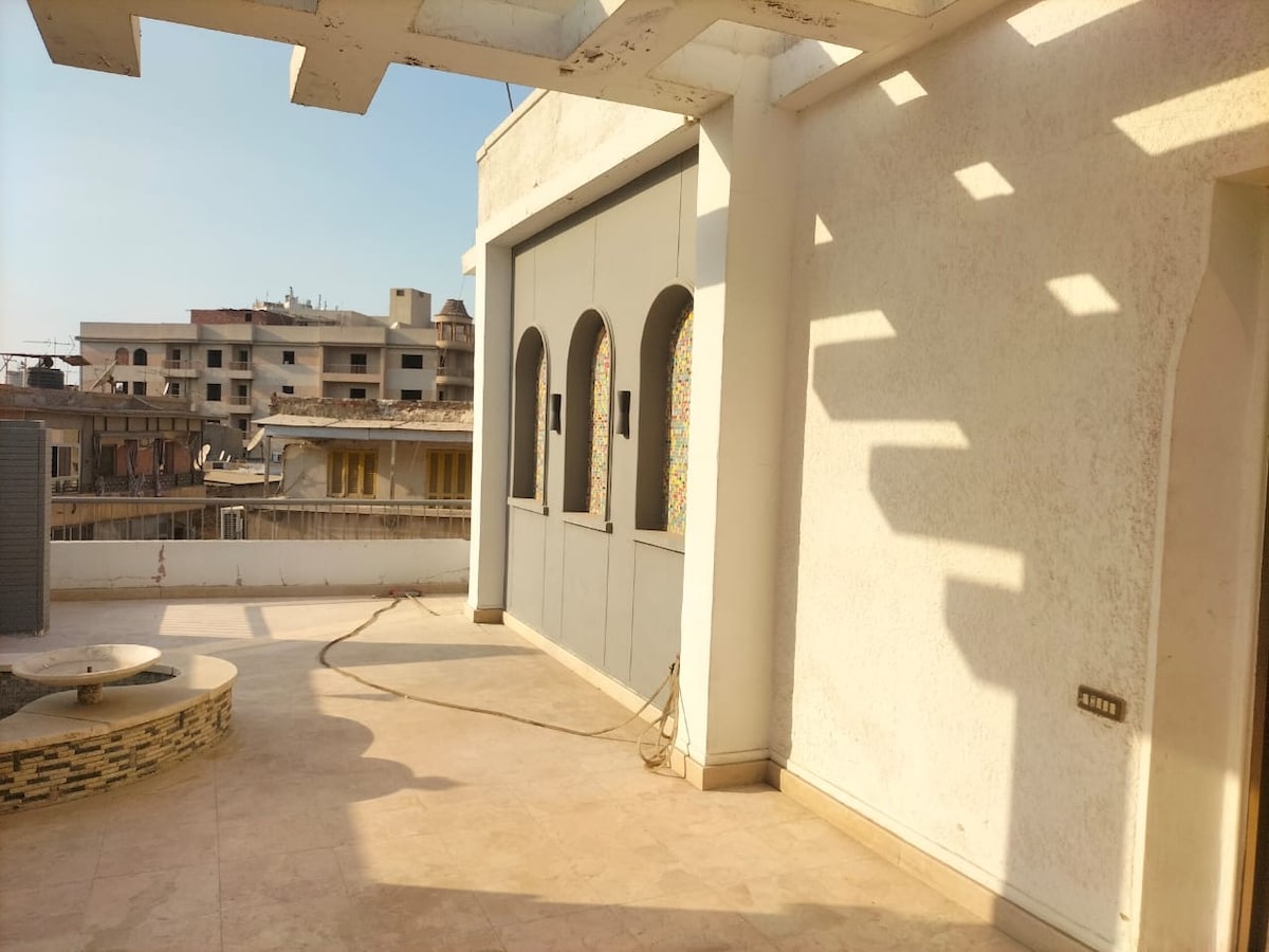 A spacious private terrace is visible, with light-colored tiles and decorative archways along one wall. Natural light casts distinctive shadows on the surface. The background features a mix of buildings, indicating a central urban location.