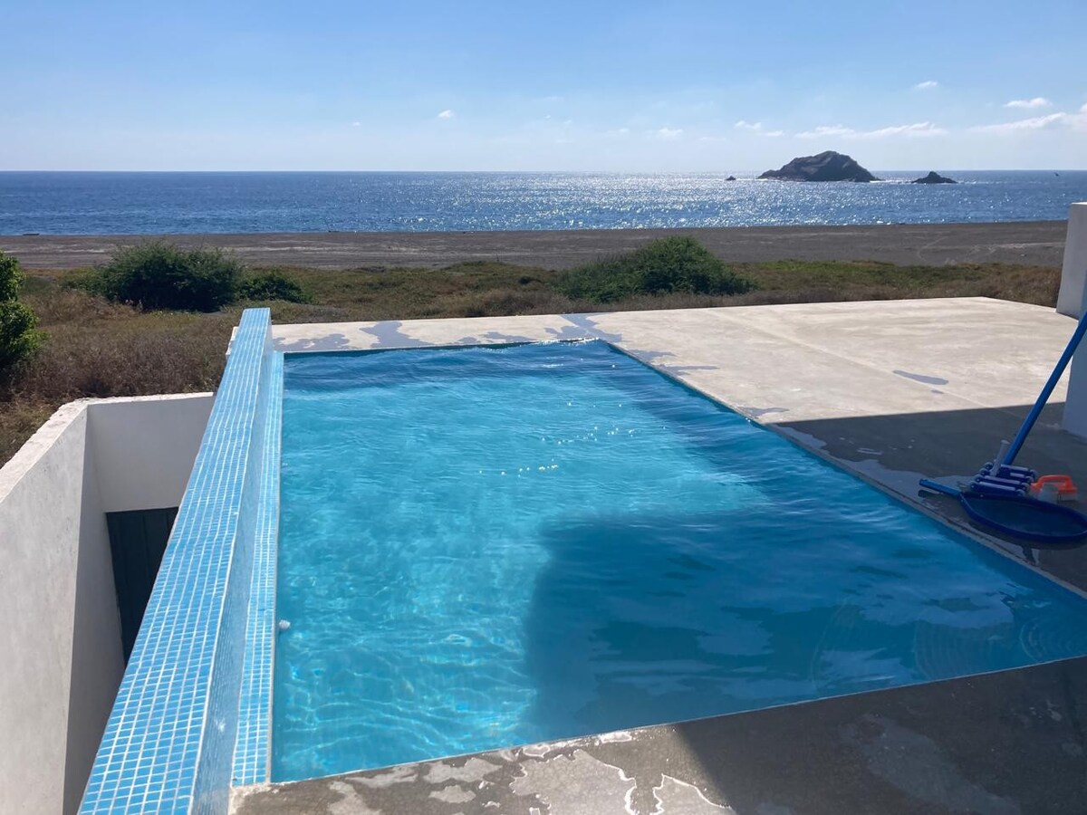 An infinity pool is positioned at the edge of a spacious terrace, offering unobstructed views of the ocean. The water reflects sunlight, while a gentle shoreline and island are visible in the distance. Surrounding greenery complements the serene landscape.