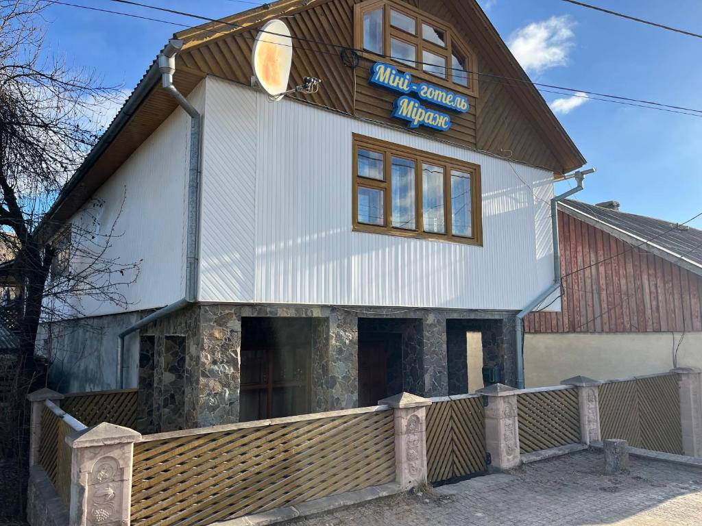 A multi-story building features a wooden sign displaying the name 'Міні-готель Міраж'. The stone and wood exterior is complemented by large windows that allow natural light to enter. A fence surrounds the property, providing a welcoming entrance.