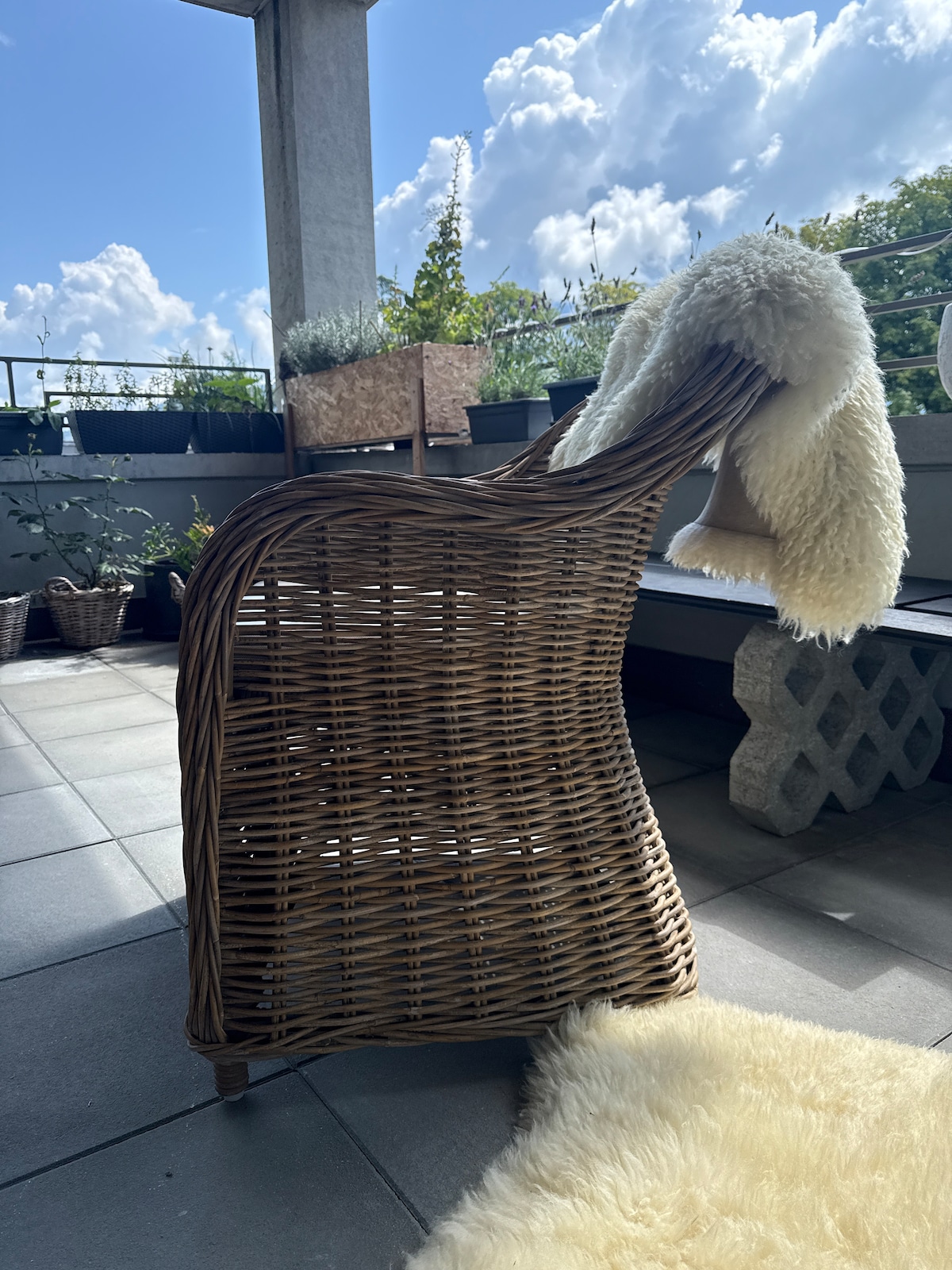 A woven wicker chair is positioned on a sunny balcony. Its curved design is complemented by a soft, cream-colored throw draped over the back. In the background, planters filled with greenery are visible under a bright blue sky with fluffy white clouds.