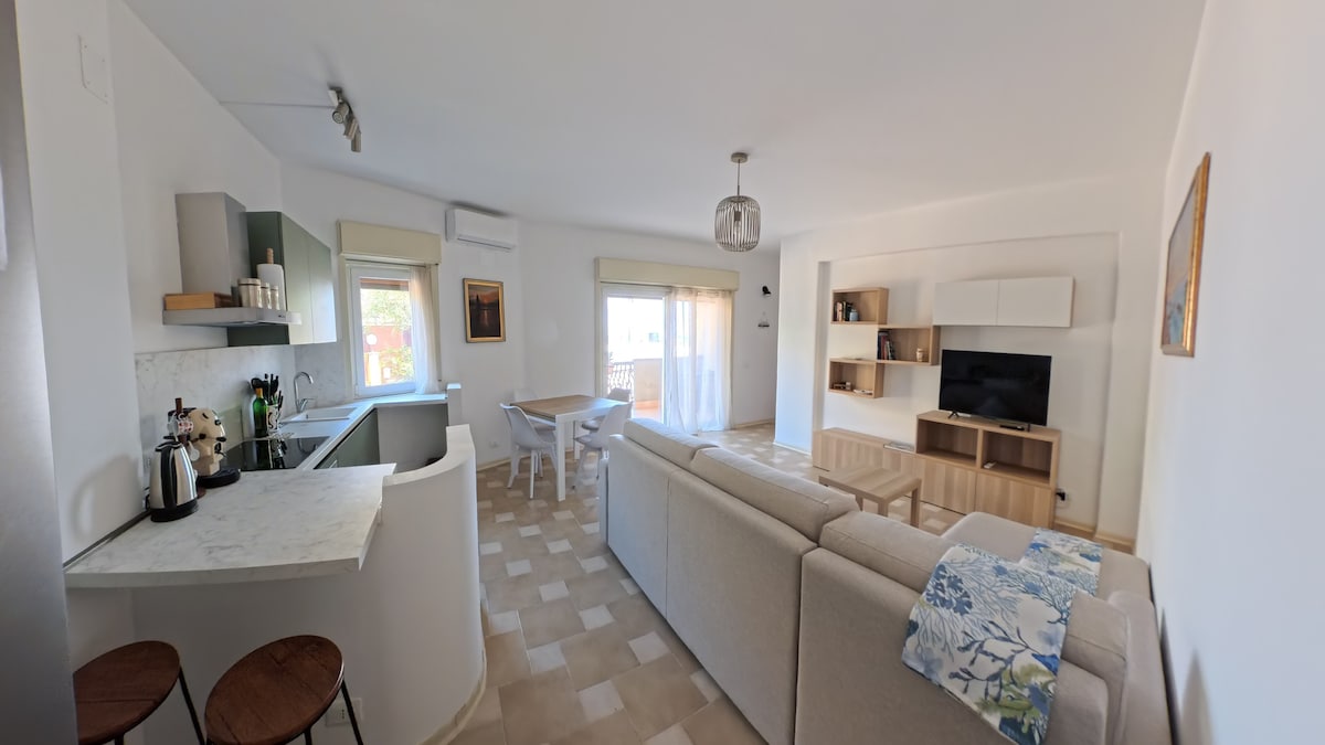 An open-plan living space is revealed, featuring a light-colored sofa centered in the room. A dining table and four chairs are visible, along with a well-equipped kitchen area that includes modern appliances. Large windows allow for ample natural light, enhancing the welcoming ambiance.