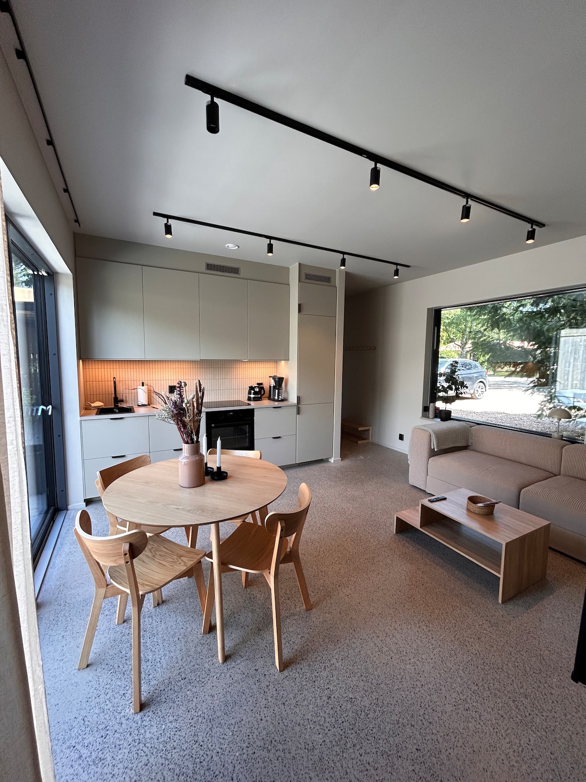 An open living and dining area features a round wooden table surrounded by four light-colored chairs. A comfortable sofa is positioned adjacent to large glass doors, creating a bright space. A compact kitchen area with modern appliances is visible in the background.