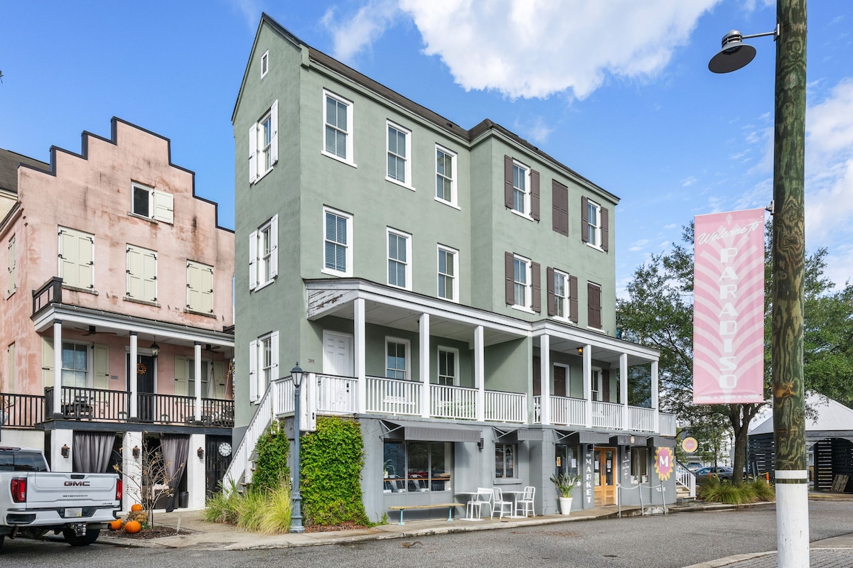 A three-story building with a green facade, featuring multiple windows and dark shutters. A covered porch spans the front, with tables and chairs arranged for outside seating. Two adjacent buildings with distinct colors complete the charming street view.