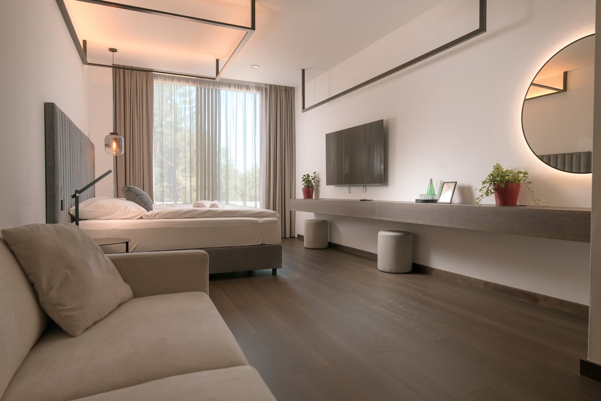 A modern guest room features a spacious bed with a cozy, neutral-colored sofa nearby. A large window allows natural light to illuminate the space, while minimalistic decor includes a round mirror and simple plant arrangements on a flat-screen TV stand.