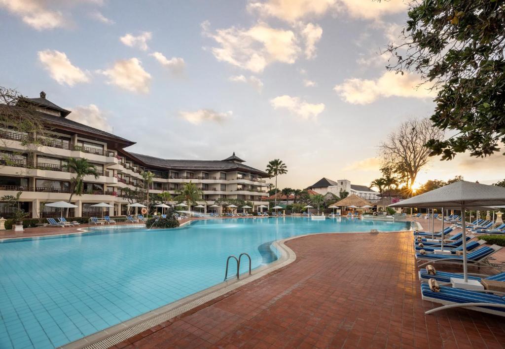 The expansive Lagoon Pool features inviting blue water, lined with comfortable lounge chairs and shaded umbrellas. Surrounding greenery and palm trees add a tropical feel, while the hotel’s architecture is visible in the background against a softly lit sky.
