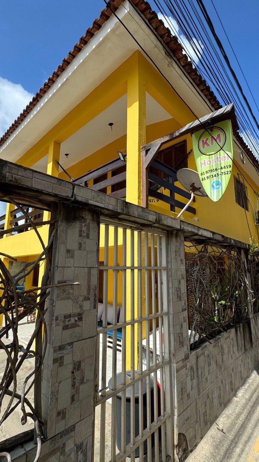 A vibrant yellow building is presented, featuring a covered balcony with wooden railings. The entrance is framed by a gated fence, with plants visible along the sides. The building's second floor is highlighted under a clear blue sky.