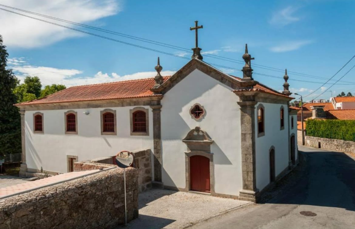 A historic church is captured from an elevated angle, showcasing its white exterior with ornate stone detailing. A red door and decorative windows add character, while a cross adorns the steeple. Surrounding greenery and a blue sky enhance the serene setting.