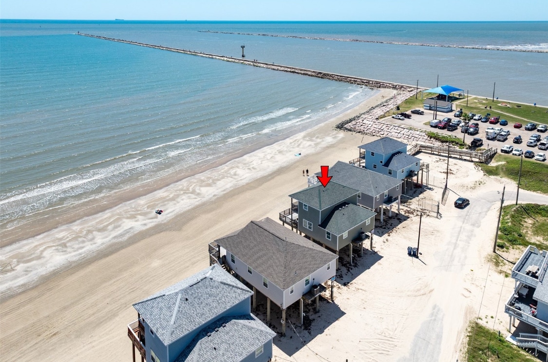 An aerial view captures the beachside location of the getaway. The sandy shore stretches along the coastline, while a jetty extends into the water. A cluster of homes is visible, with one highlighted, alongside a parking area and green space in the distance.