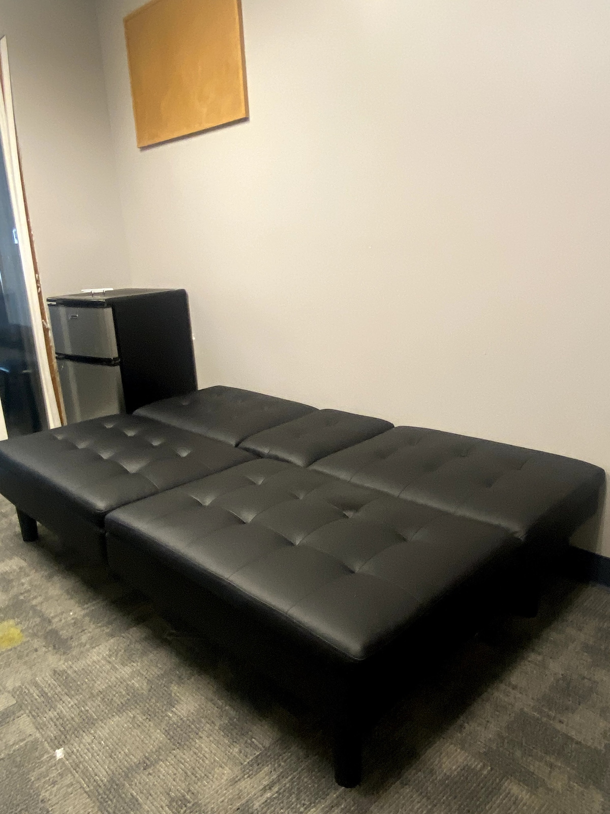 An upholstered black sofa is situated against a light grey wall, featuring a tufted design. A compact fridge is located nearby, and the flooring displays a muted patterned carpet.
