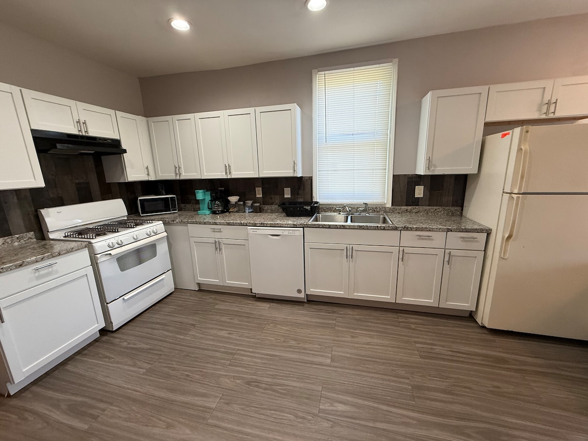 A modern kitchen showcases white cabinetry contrasted by gray granite countertops. Stainless steel appliances, including a gas stove, dishwasher, and refrigerator, are present. Natural light floods the space through a large window, enhancing the clean and functional layout.