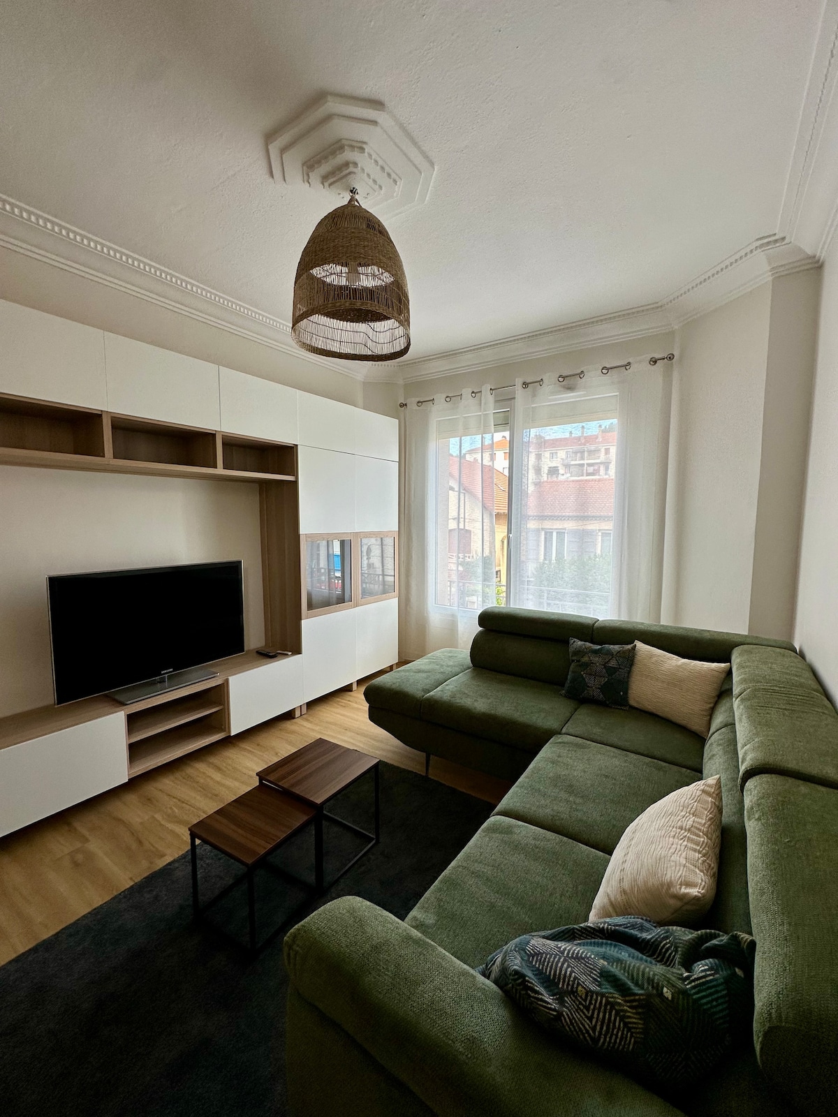 A modern living area features a spacious green sectional sofa, accented with decorative pillows. A flat-screen television is mounted on a light-colored media console. Large windows allow natural light to filter in, with sheer curtains softly diffusing the sunlight.