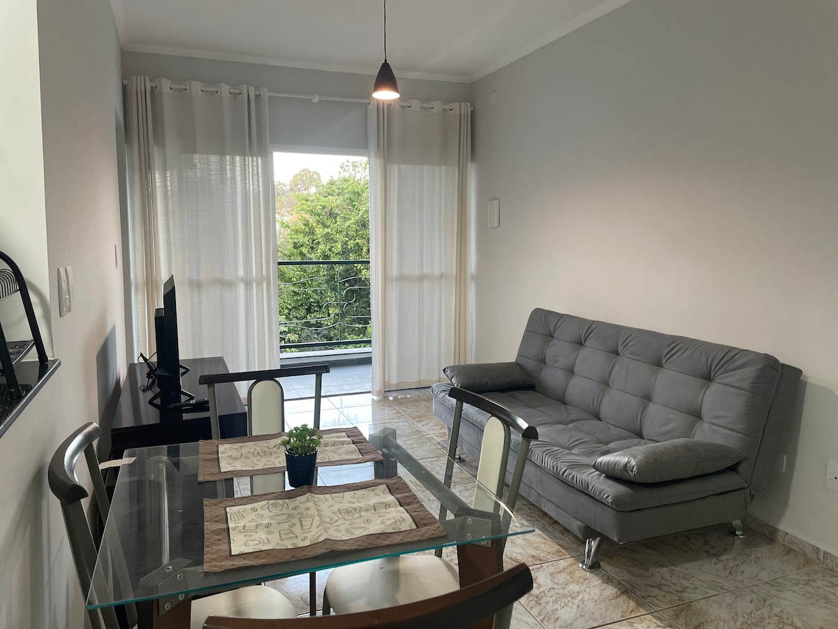 A light-filled living area features a gray sofa bed and a glass dining table set for two. Sheer curtains frame the windows, offering a view of greenery outside. A small plant sits centrally on the table, enhancing the minimalist decor.