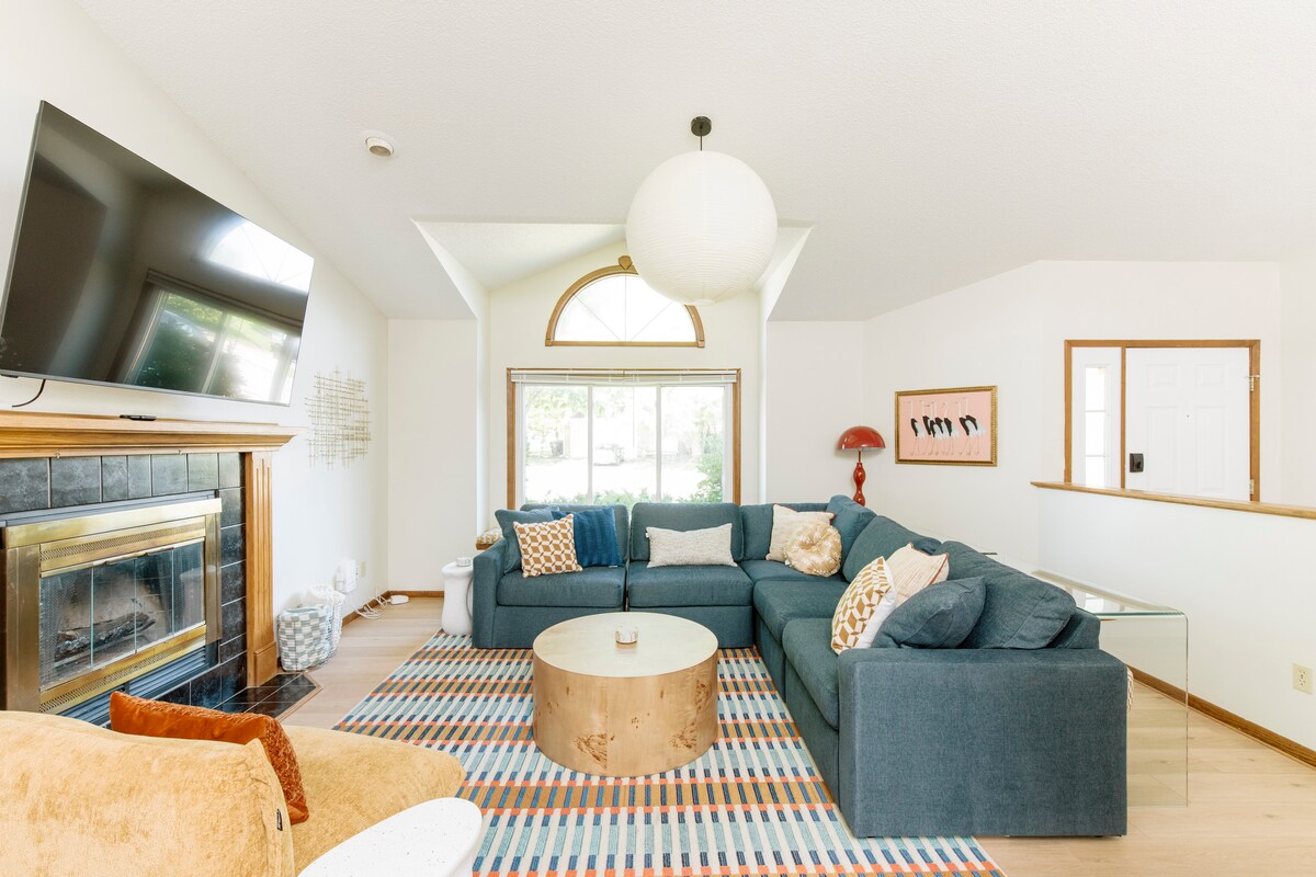 A light-filled living room showcases a large sectional sofa in deep blue, arranged around a circular coffee table. A flat-screen TV is mounted above a black fireplace, with natural light entering through a prominent arched window. A bold area rug adds texture and warmth to the space.