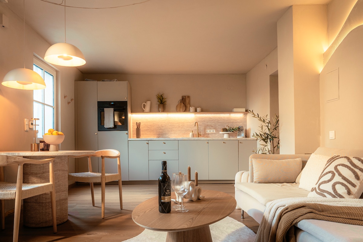 An open living space is showcased, featuring a modern kitchen with light-colored cabinetry and a dining area with two woven chairs. A cozy lounge area includes a light sofa and a wooden coffee table, complemented by soft lighting that creates a warm ambiance.