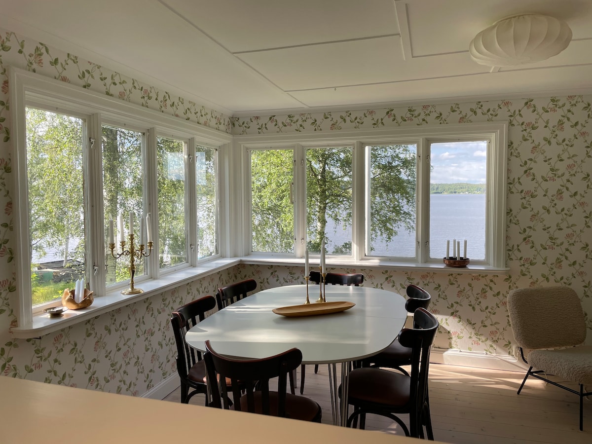 A bright dining area is surrounded by large windows, offering views of the lake and greenery outside. The table accommodates multiple chairs and is complemented by a decorative centerpiece. Soft floral wallpaper adds a touch of warmth to the space, enhancing the serene atmosphere.
