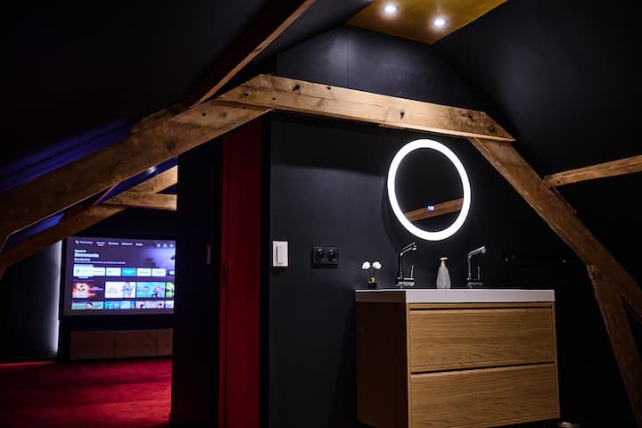 Uno spazio bagno essenziale ed elegante, tra il nero intenso e il calore del legno. Lo specchio luminoso rotondo ricorda le luci di un set cinematografico: atmosfera garantita.