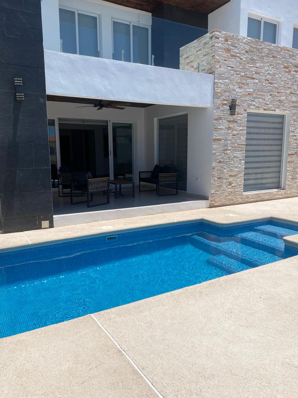 A crystal-clear pool is set in a smooth patio area, bordered by modern stone and stucco walls. Comfortable outdoor seating is visible in a shaded area beyond the pool, with large sliding glass doors leading inside. Sunlight reflects off the water, creating a serene atmosphere.