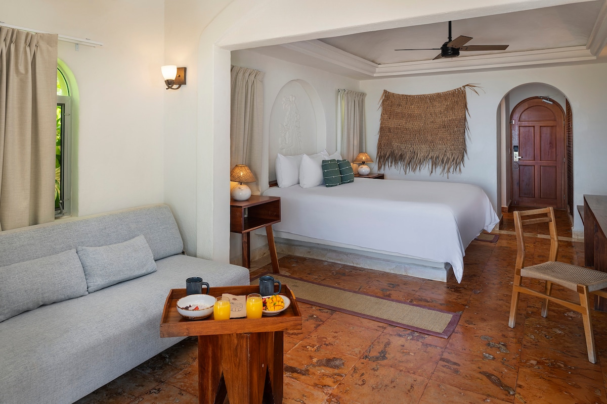 A spacious room features a comfortable seating area with a light gray sofa and a wooden table set with breakfast items. A large bed dominates the space, complemented by soft lighting from bedside lamps. Natural textures are introduced by woven wall decor and warm earth-toned tiles.